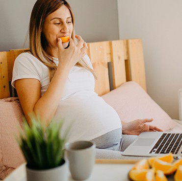 Pregnant woman eating a slice of orange working on laptop